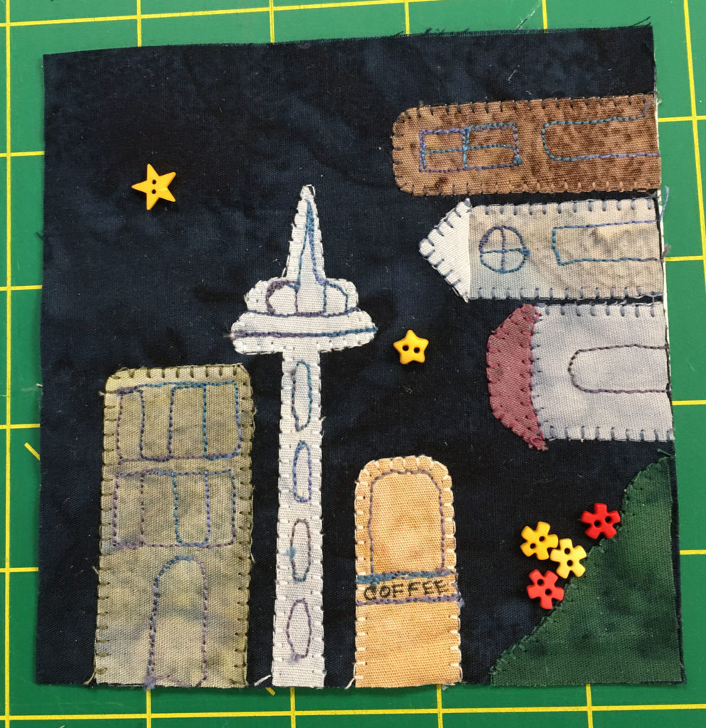 This quilt block features building going around a corner with buttons depicting flowers in the bottom right. The space needle, a tall building with a particular shape, sits next to a coffee shop.