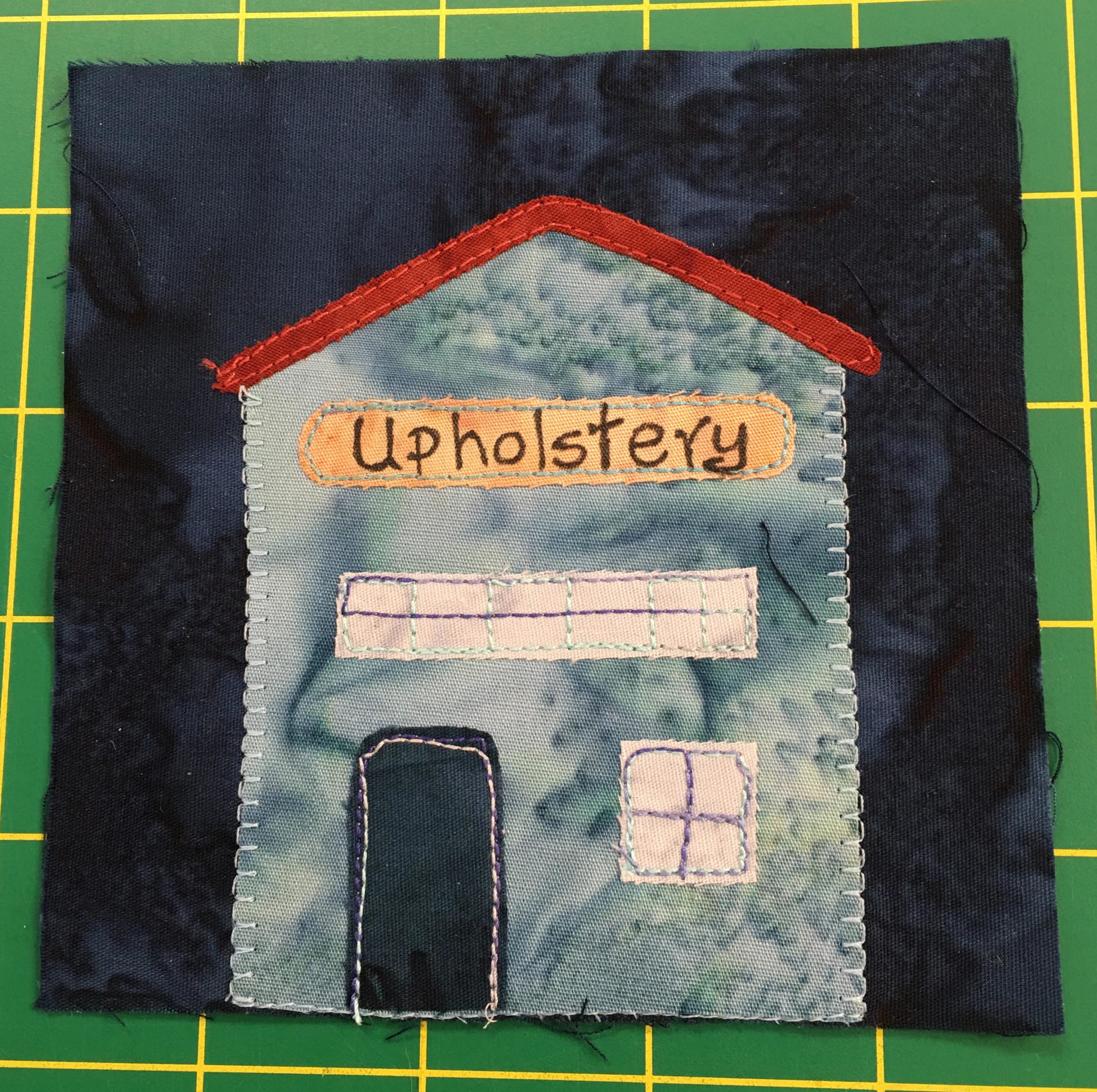 A quilt block with a tall gray building against a dark background indicating night. On the front of the building above the dark door is a sign reading 