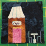 This quilt lock shows a caramel colored café with a pink door. Above the pink door it reads