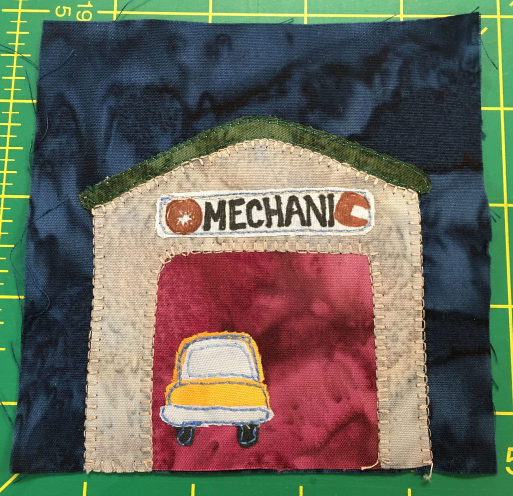 This quilt block shows a garage against a dark background. The inside of the garage is a rich red and an orange car is inside.