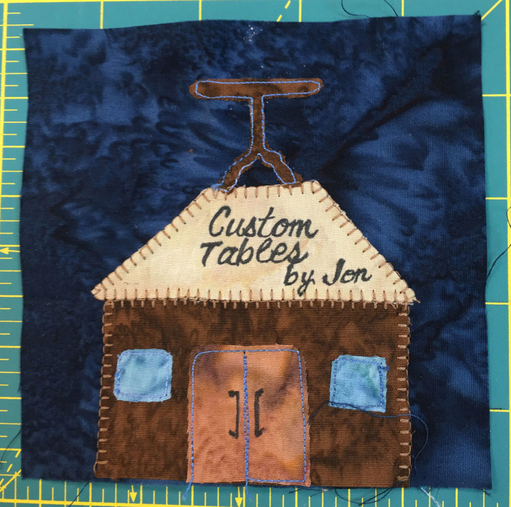 This quilt block depicts a building with a table on top. The sign on the front reads