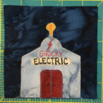 This quilt block shows a gray metal building against a dark background. It has red double doors, and a lightbulb on the top of it. The front reads,