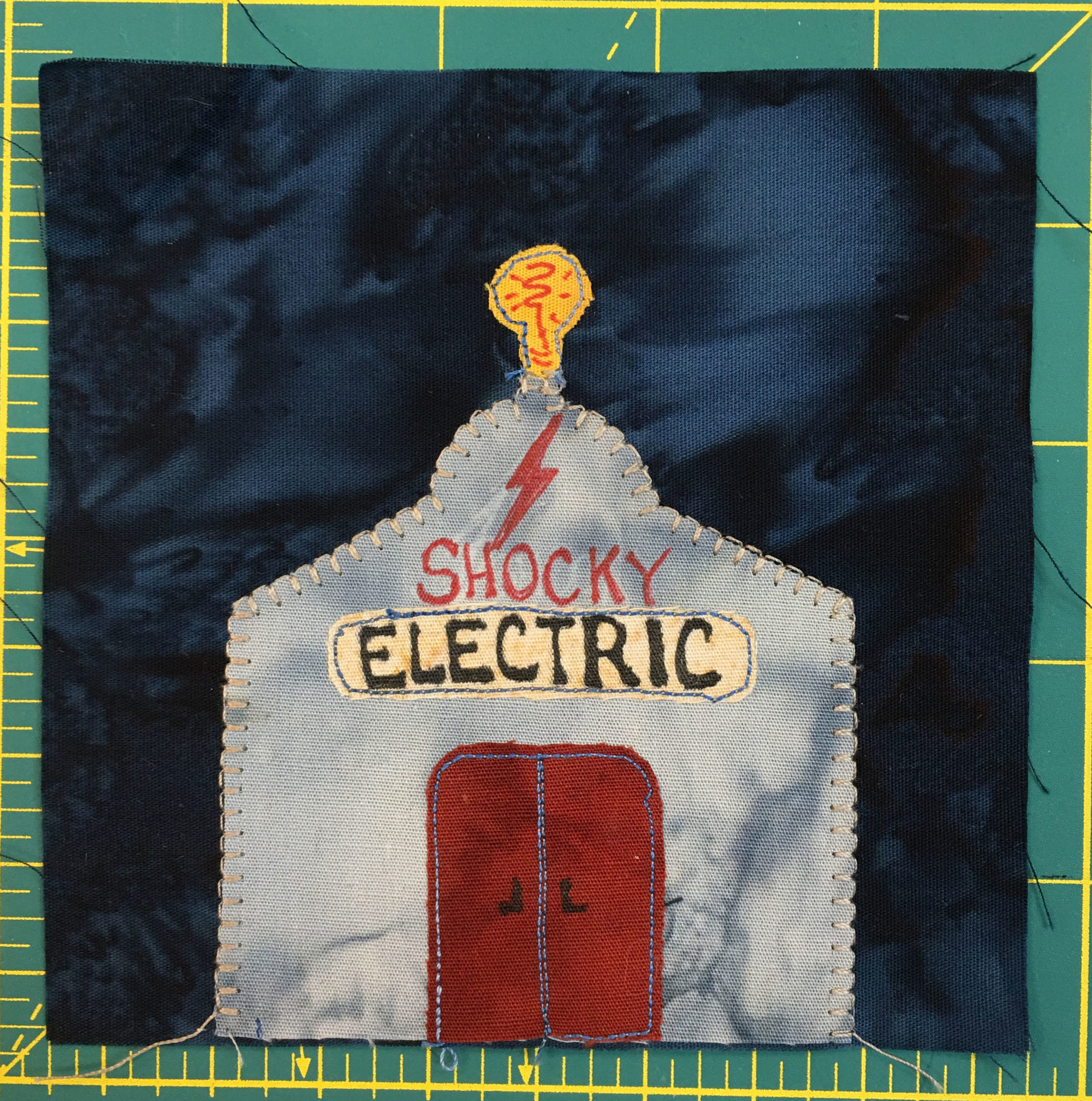 This quilt block shows a gray metal building against a dark background. It has red double doors, and a lightbulb on the top of it. The front reads,