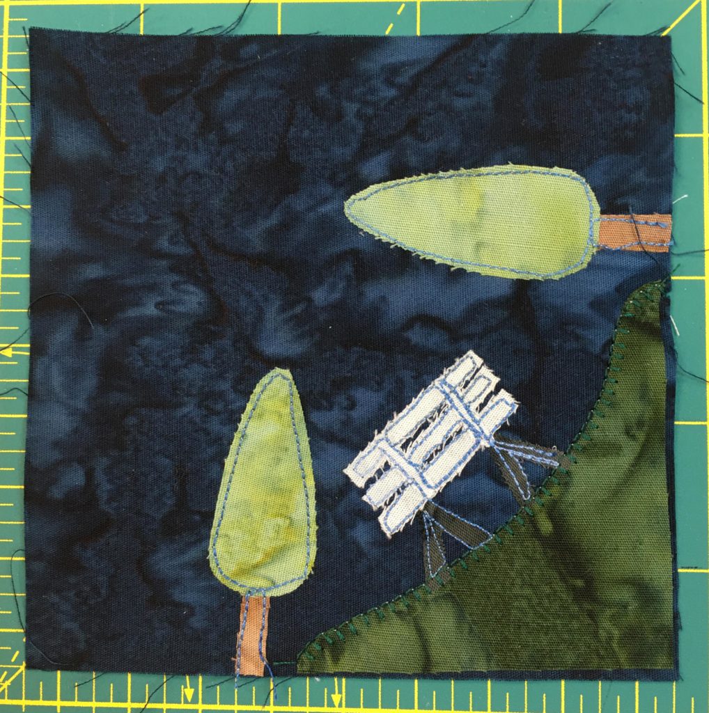 This quilt block shows a patch of grass in the bottom right with a white bench and two trees, one vertical and one horizontal. The patch of grass overlaps the corner on the botom and right sides.