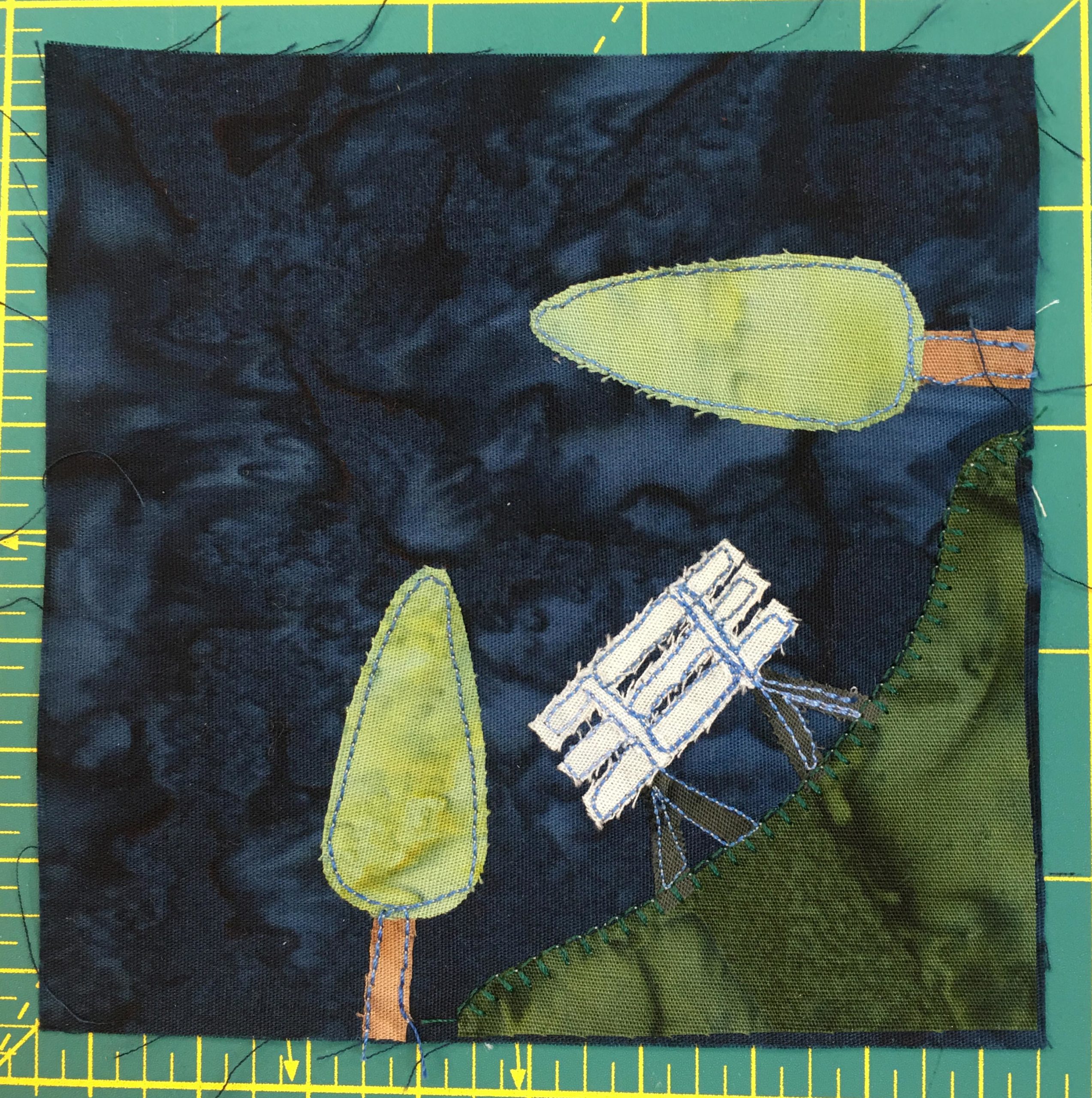 This quilt block shows a patch of grass in the bottom right with a white bench and two trees, one vertical and one horizontal. The patch of grass overlaps the corner on the botom and right sides.