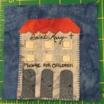 This quilt shows a tall gray building with a red roof. It has three doors and six windows in two horizontal rows. The words on the front read,