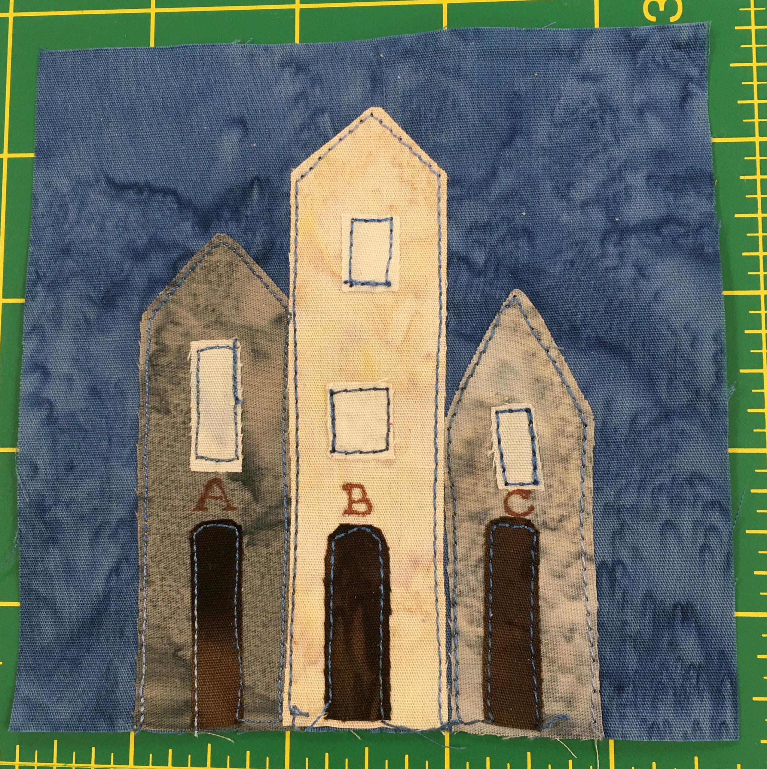 This quilt block shows three tall buildings with pointy roofs. The middle lighter one is two stories tall. They are labeld above the dark doors with the letters A, B, and C.
