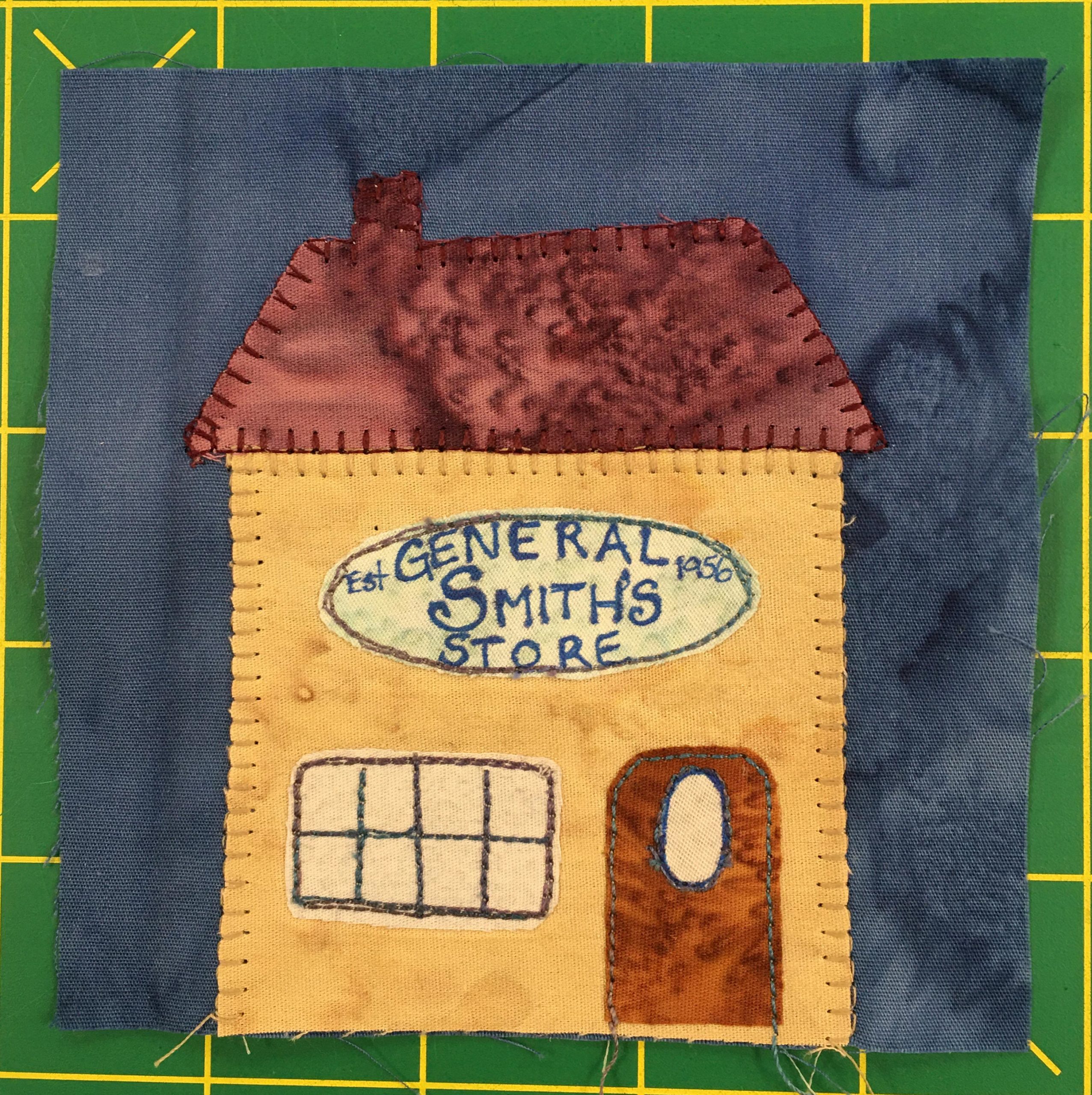 This quilt block shows a yellow house with a deep red roof and chimney. On the front a sign reads, 