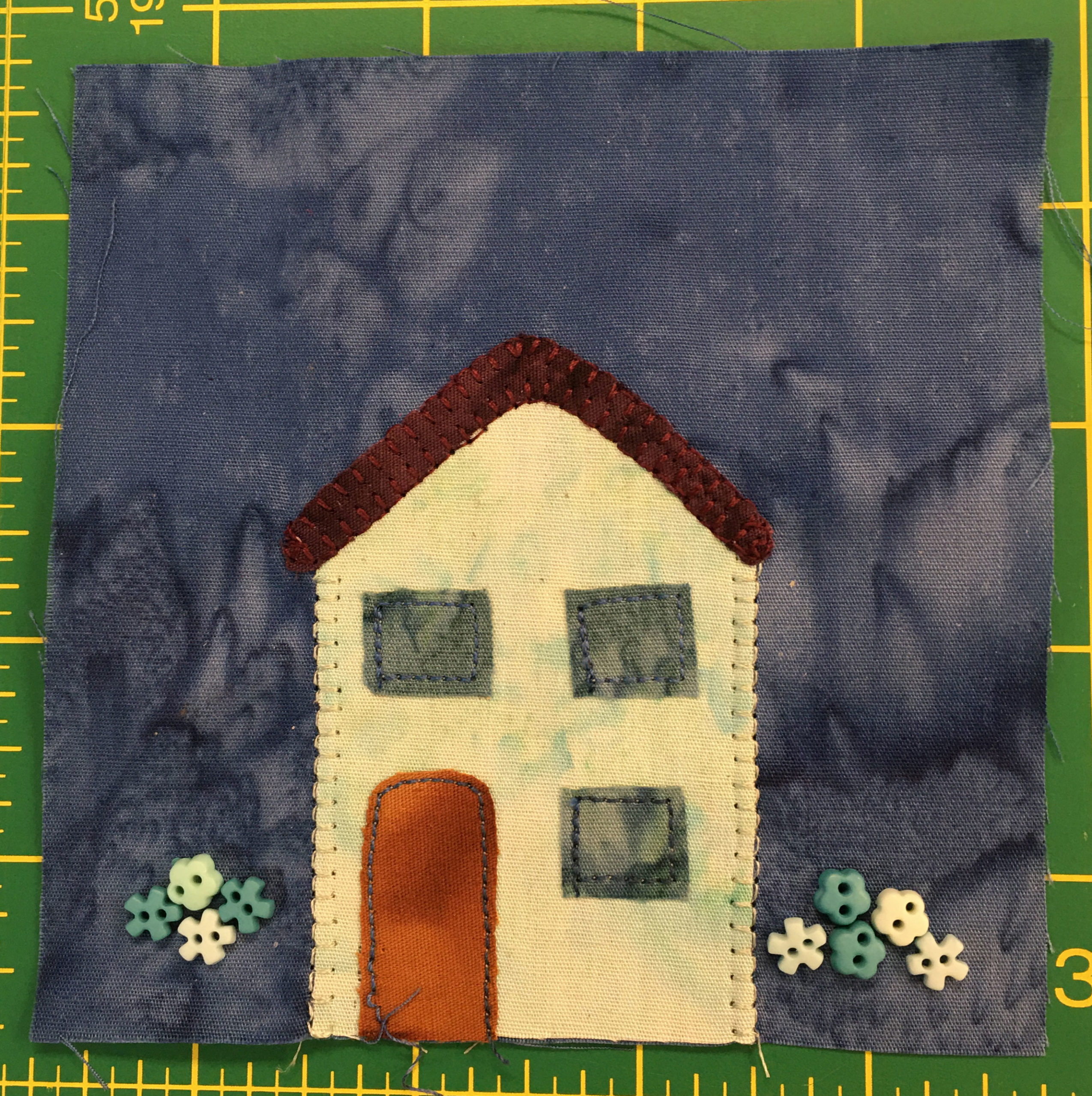 This quilt block shows a straight forward house with three windows and a red door. The house is off white. Next to the house are buttons showing flower blossoms.