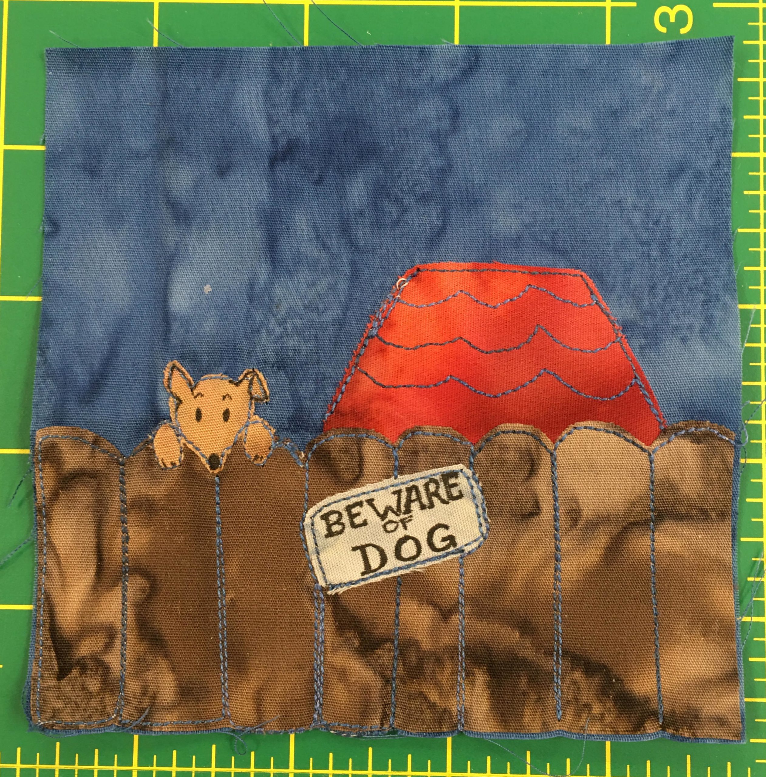 This quilt block shows a brown fence in front of a house. You can only see the red roof of the house. On the fence is a sign that says,