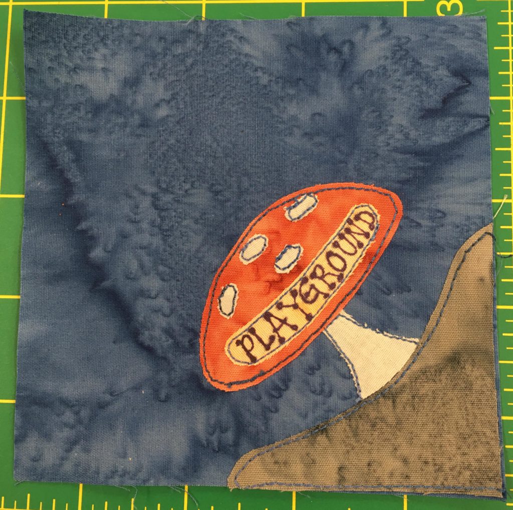 This quilt block shows corner plot. In the bottom right corner is a brown piece of ground with a large orange mushroom looking thing on top with a sign on it that reads, 