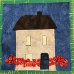 This quilt block shows a tall square house with a dark roof and a chimney. There are two floors with three windows and a dark door. All along the bottom of the square are small red buttons in various little shapes, including hearts, to signify flowers.
