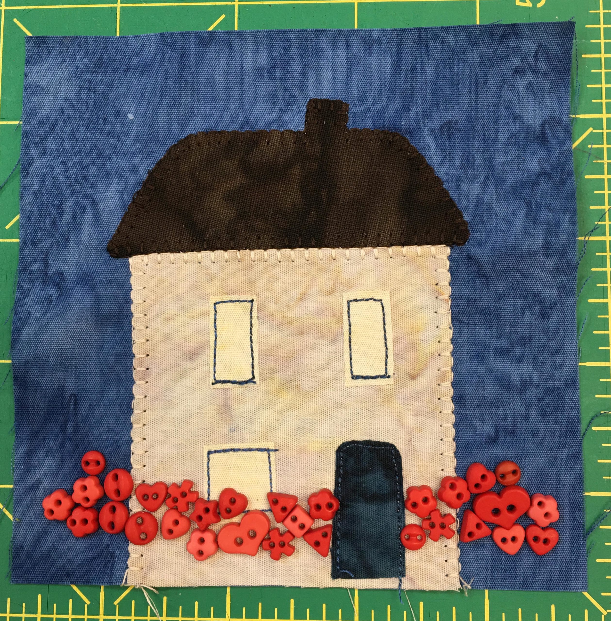 This quilt block shows a tall square house with a dark roof and a chimney. There are two floors with three windows and a dark door. All along the bottom of the square are small red buttons in various little shapes, including hearts, to signify flowers.