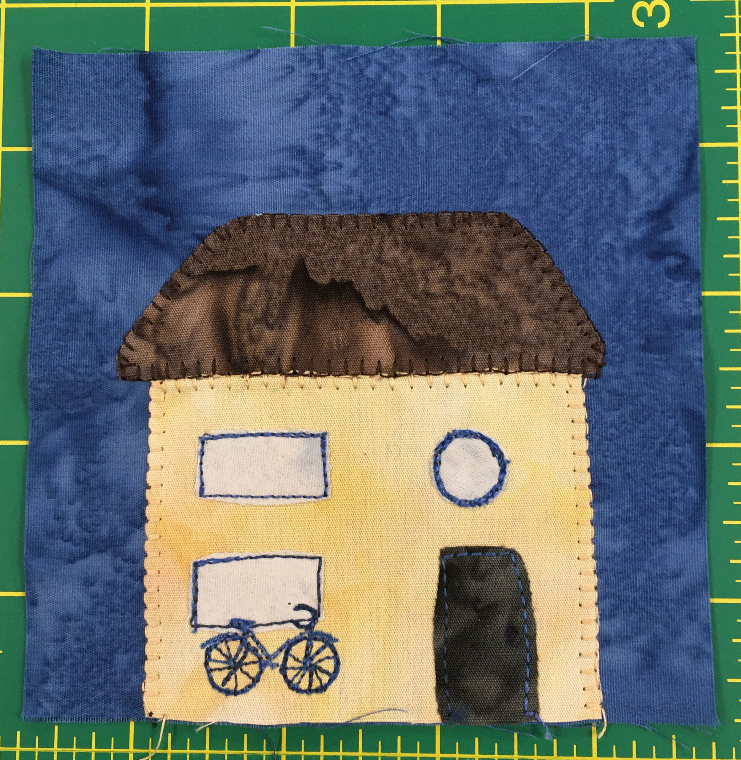 This quilt block shows a blue background and a yellow house. The house has a bicycle in front and two rectangular windows. Above the dark door on the right is a circular window.