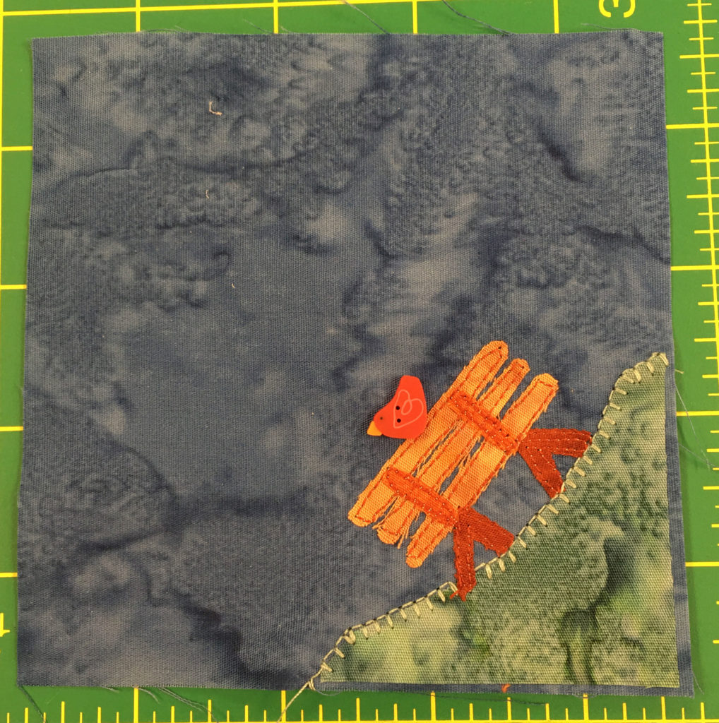 This quilt block shows a blue gray background and a patch of grass in the bottom right. On the patch of grass is an orange bench and on the the bench is a little red bird with a yellow beak facing left.