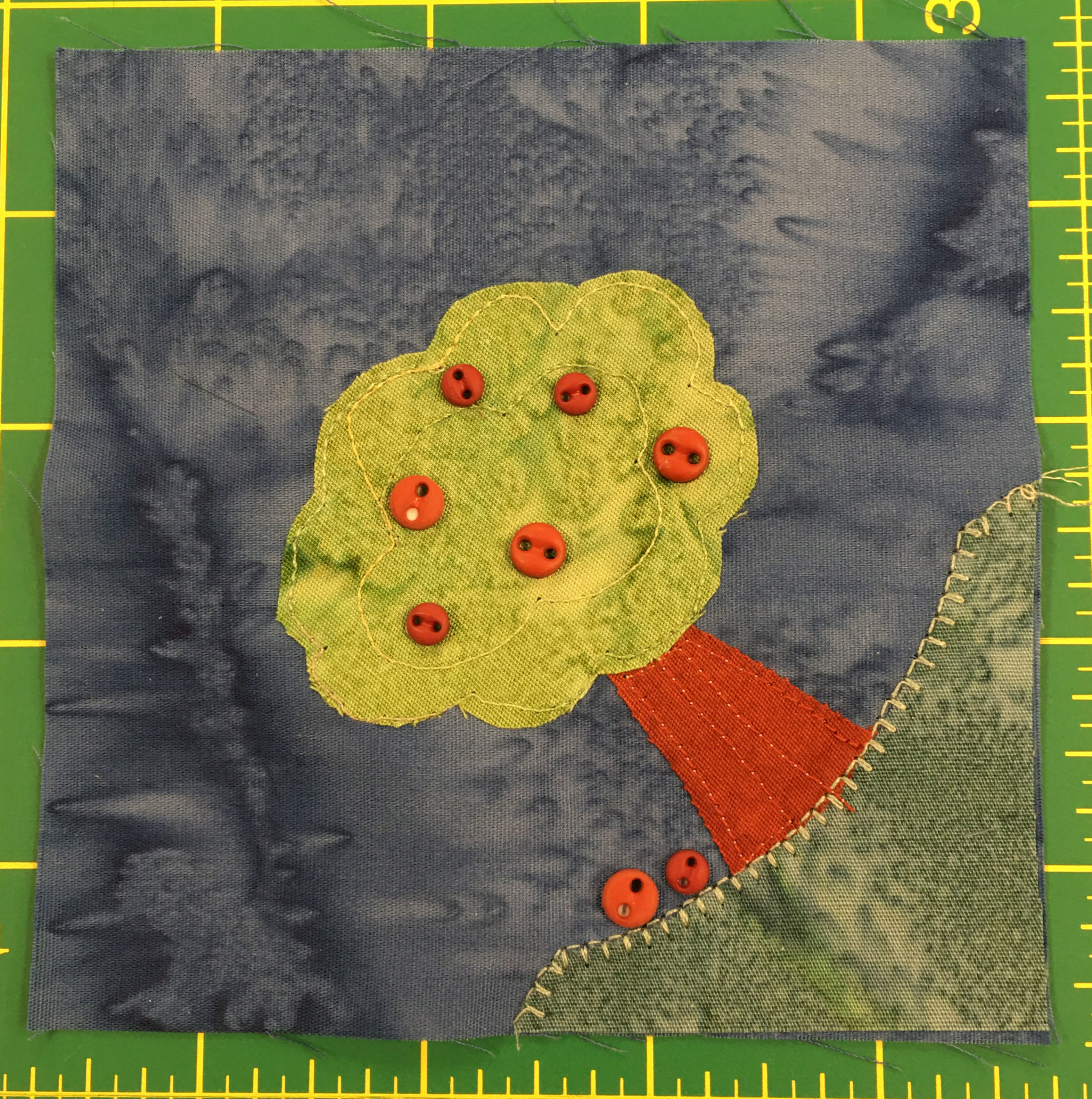 This quilt block shows a dark blue background with a path of green grass in the lower right corner. There is a big apple tree on the grass jutting out into the center of the square. The apples are actually small round red two holed buttons of varying sizes.