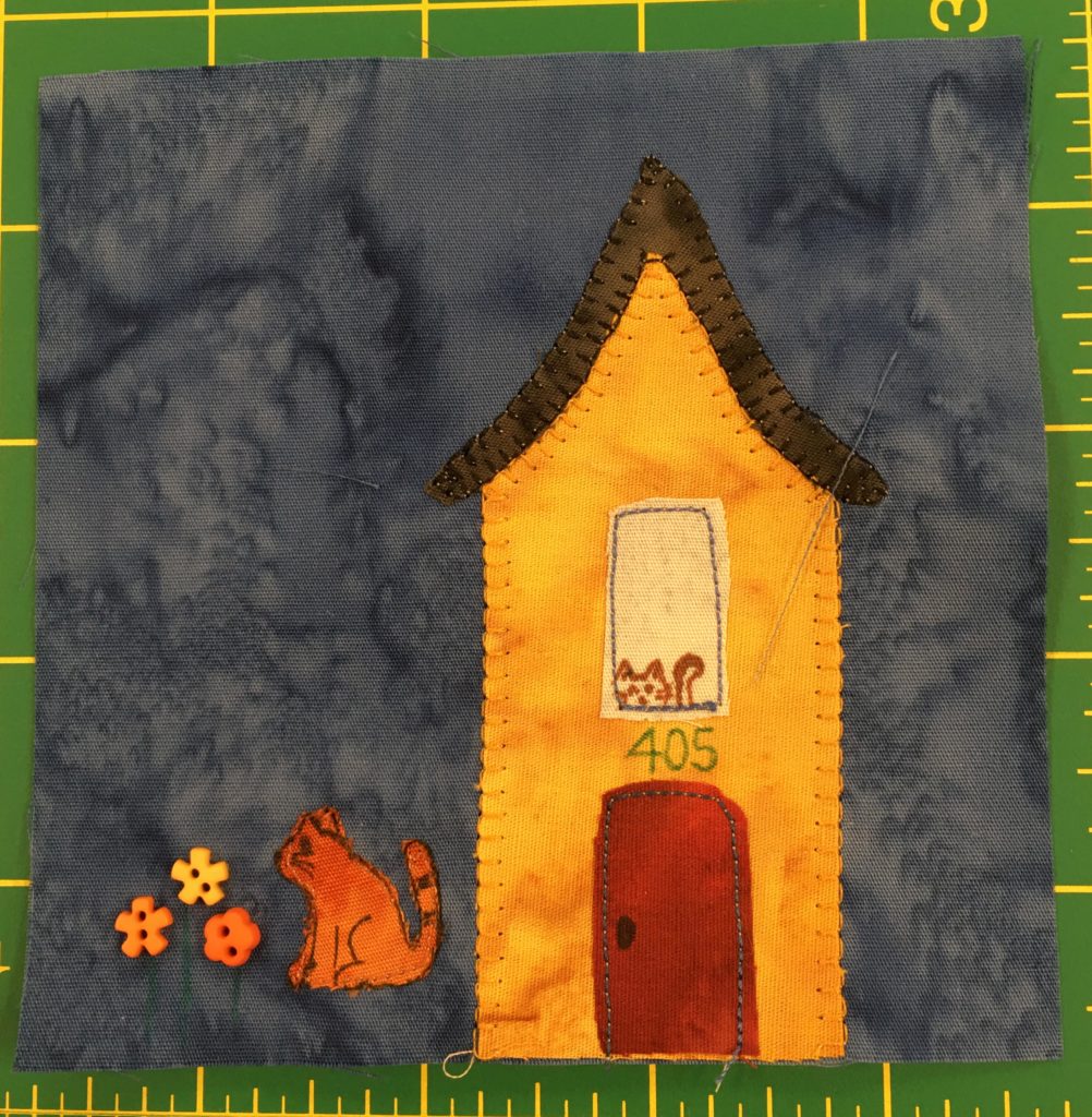 This block shows a tall yellow-orange house with a red door. In the tall window above the door you can see a cat peering out. To the left of the building you can see a cat sitting next to some flowers which are signified by some colorful fun shaped buttons.
