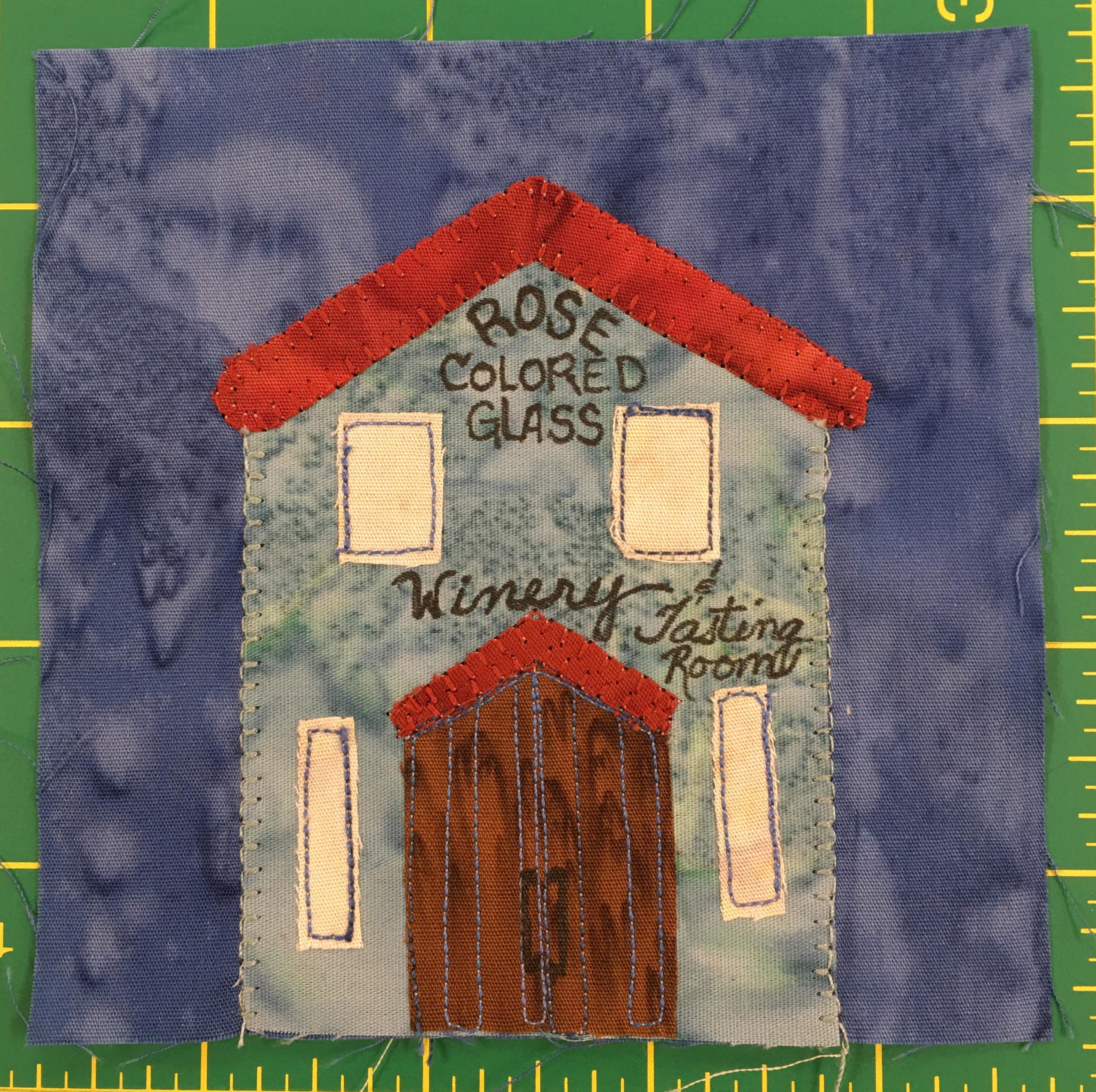 This quilt block shows a tall gray-blue building with a red triangular roof. There are two windows on the second floor and two tall windows on the first floor next to a set of arched double doors. On the building are the words,