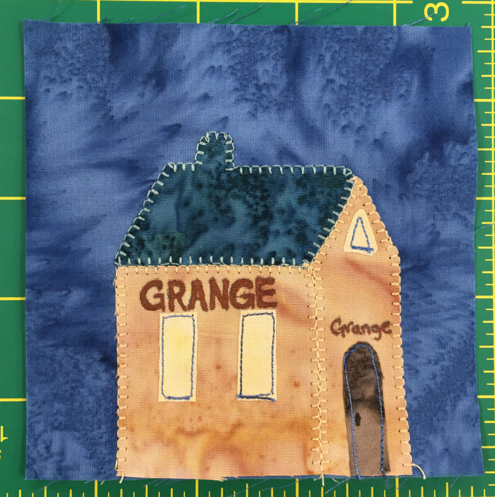 This quilt block shows an orange building from a side angle with a dark blue roof. There are two tall windows on the side and a brown door on front. Across the side and the front is the word,