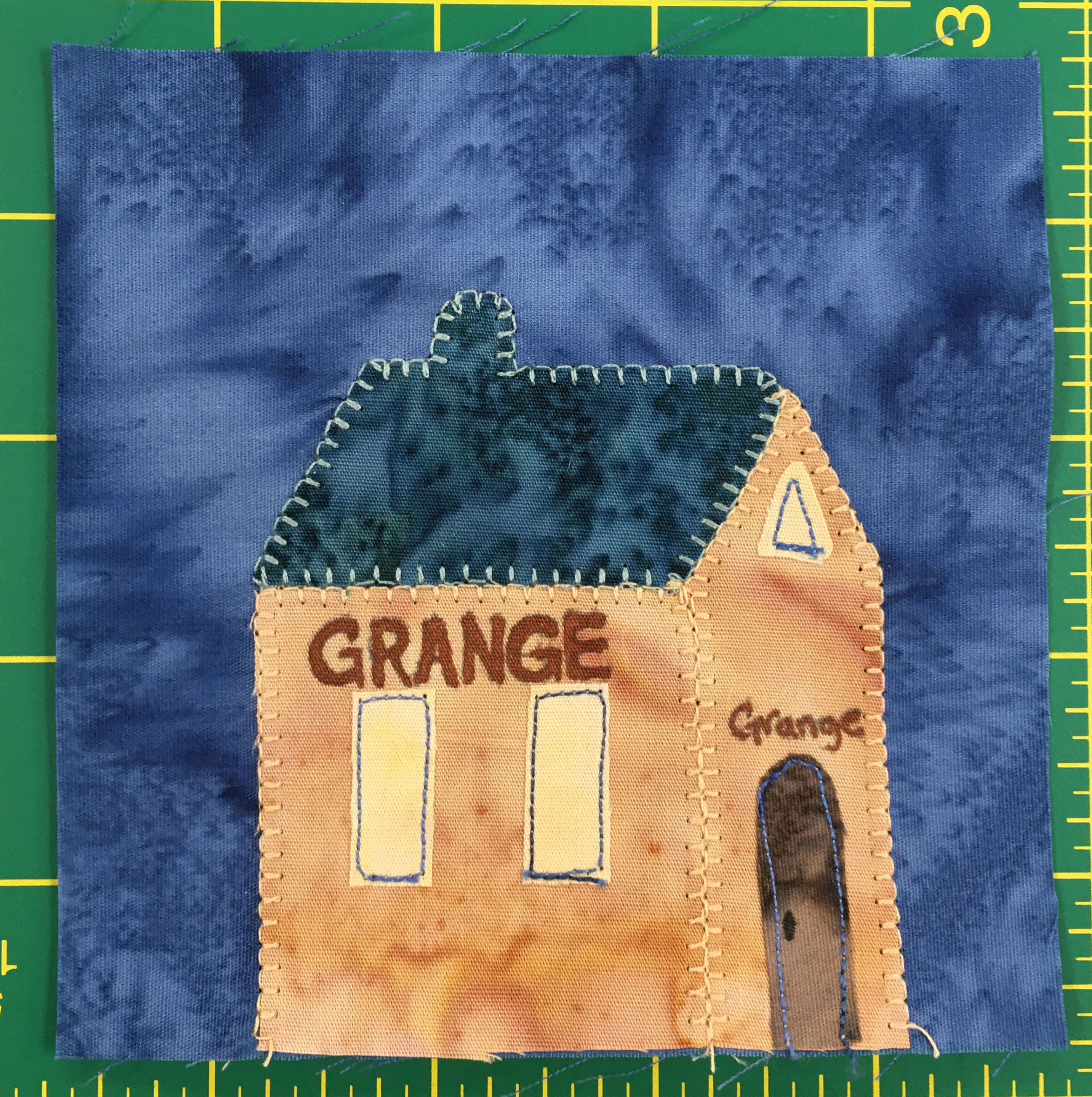 This quilt block shows an orange building from a side angle with a dark blue roof. There are two tall windows on the side and a brown door on front. Across the side and the front is the word,