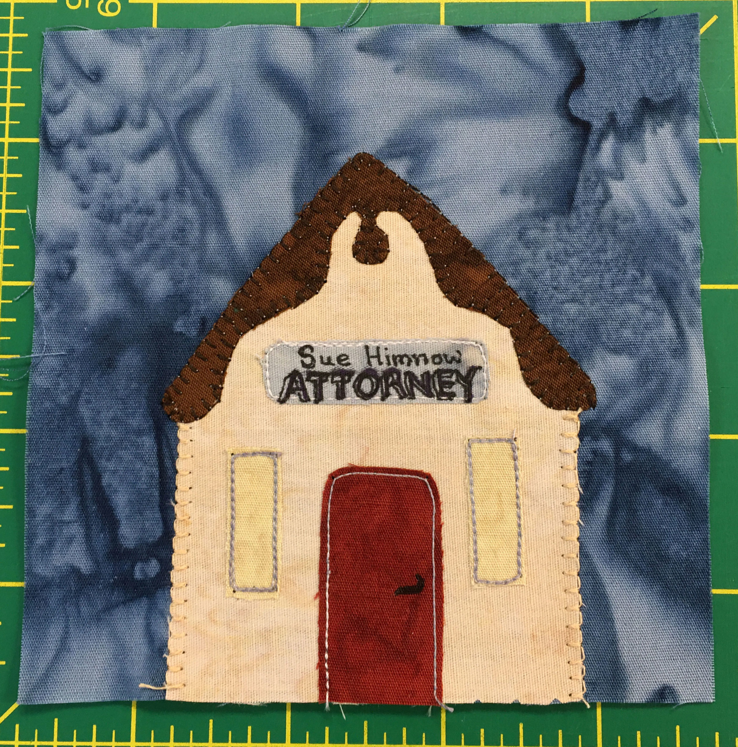 This quilt block shows the front of a triangle shaped yellow building. It has a brown roof that looks a bit like a chocolate ganache drizzled on top of a cake, and a silver sign that reads, 