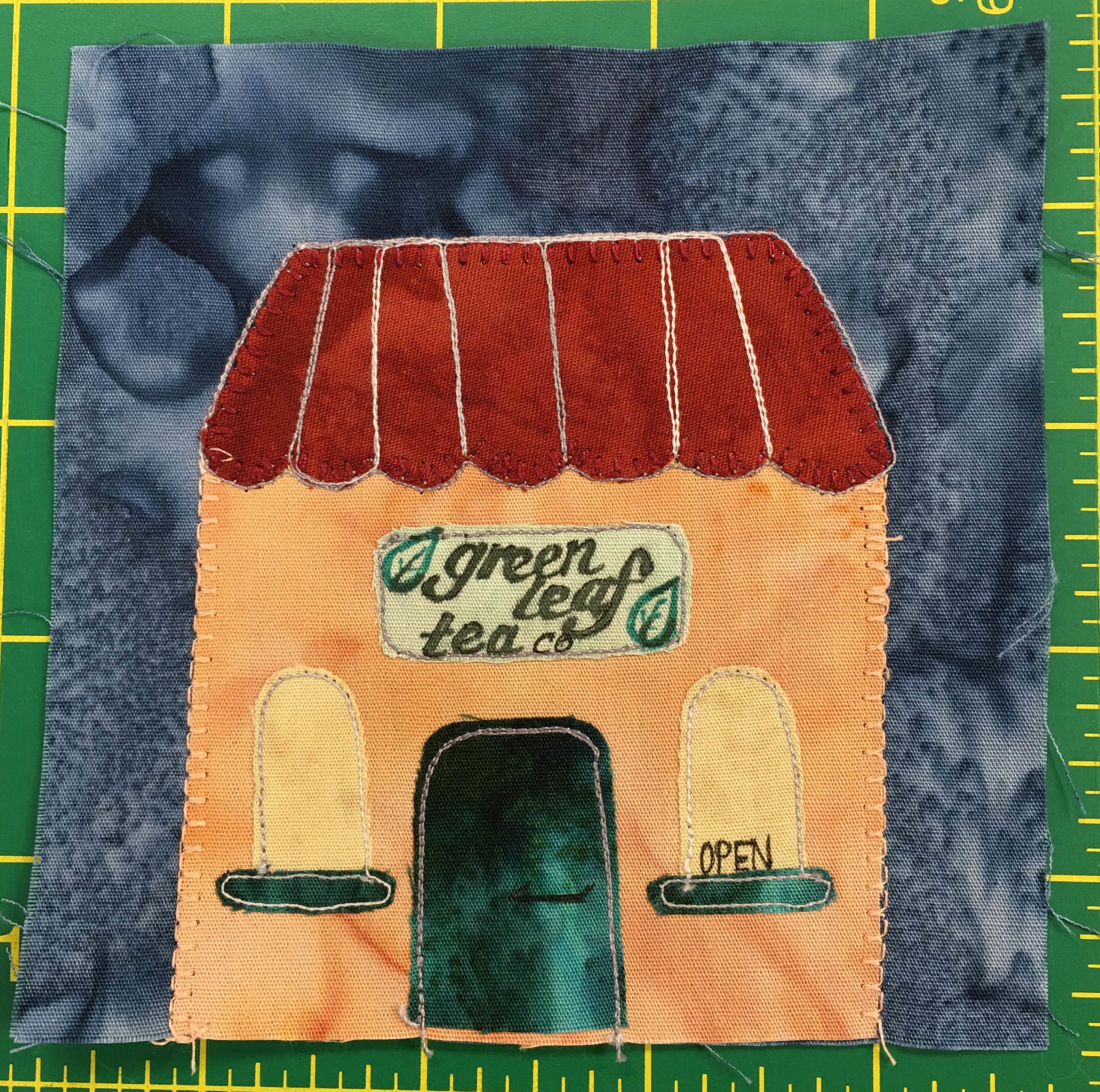 This quilt block shows an orange building with two domed yellow windows on either side of the green door. The windows are framed on the bottom by the same green trim. The roof is paneled and red. A sign with leaves on it reads,