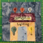 This quilt block shows a yellow building with three lightbulbs on top. There are also two large lightbulbs to either side of the big gray door. There is a sign on the square flat purple roof that reads,