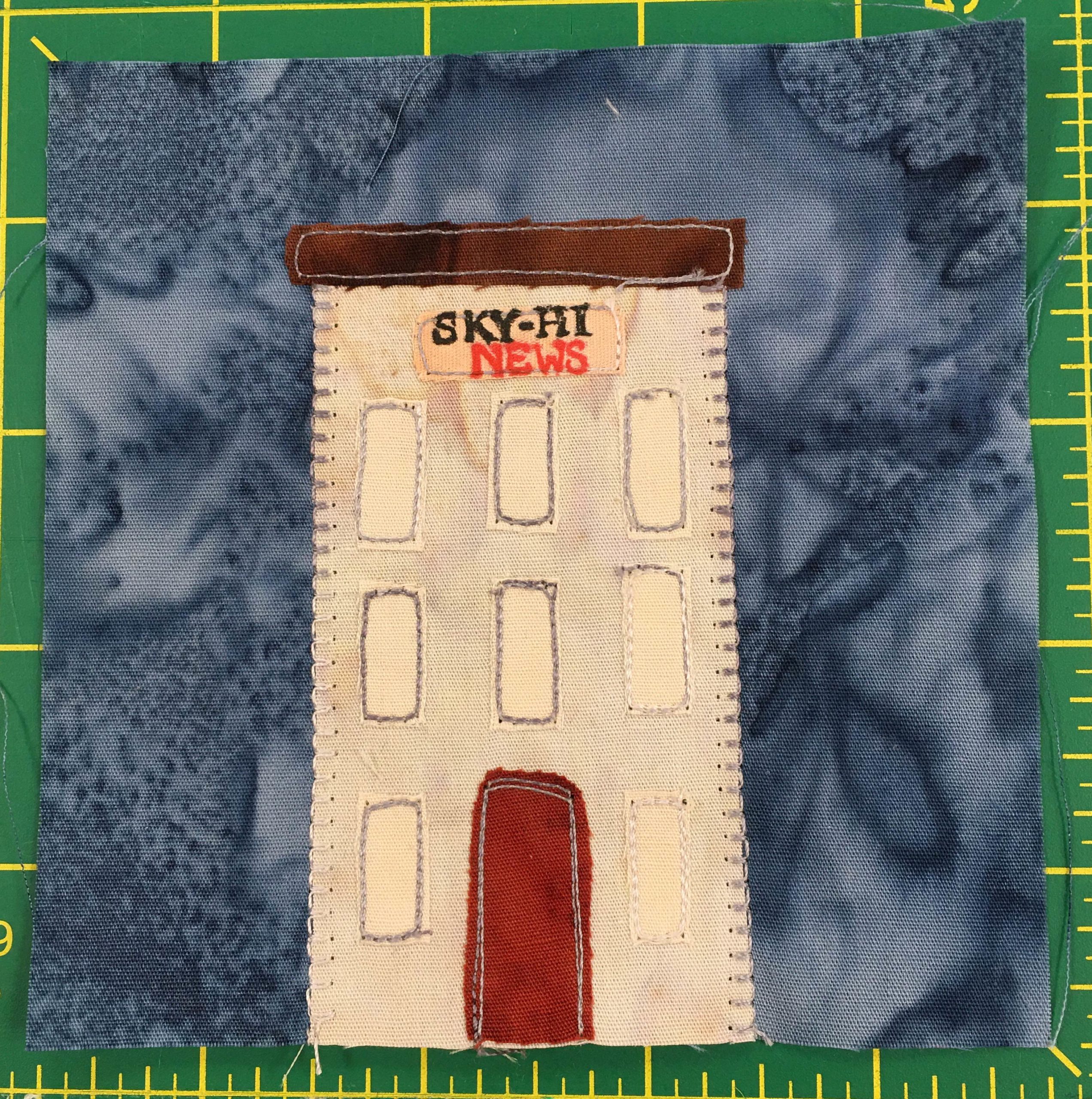 This quilt block shows a very tall white building with many tall windows and a red door. The roof is flat and brown. The front top sign reads,