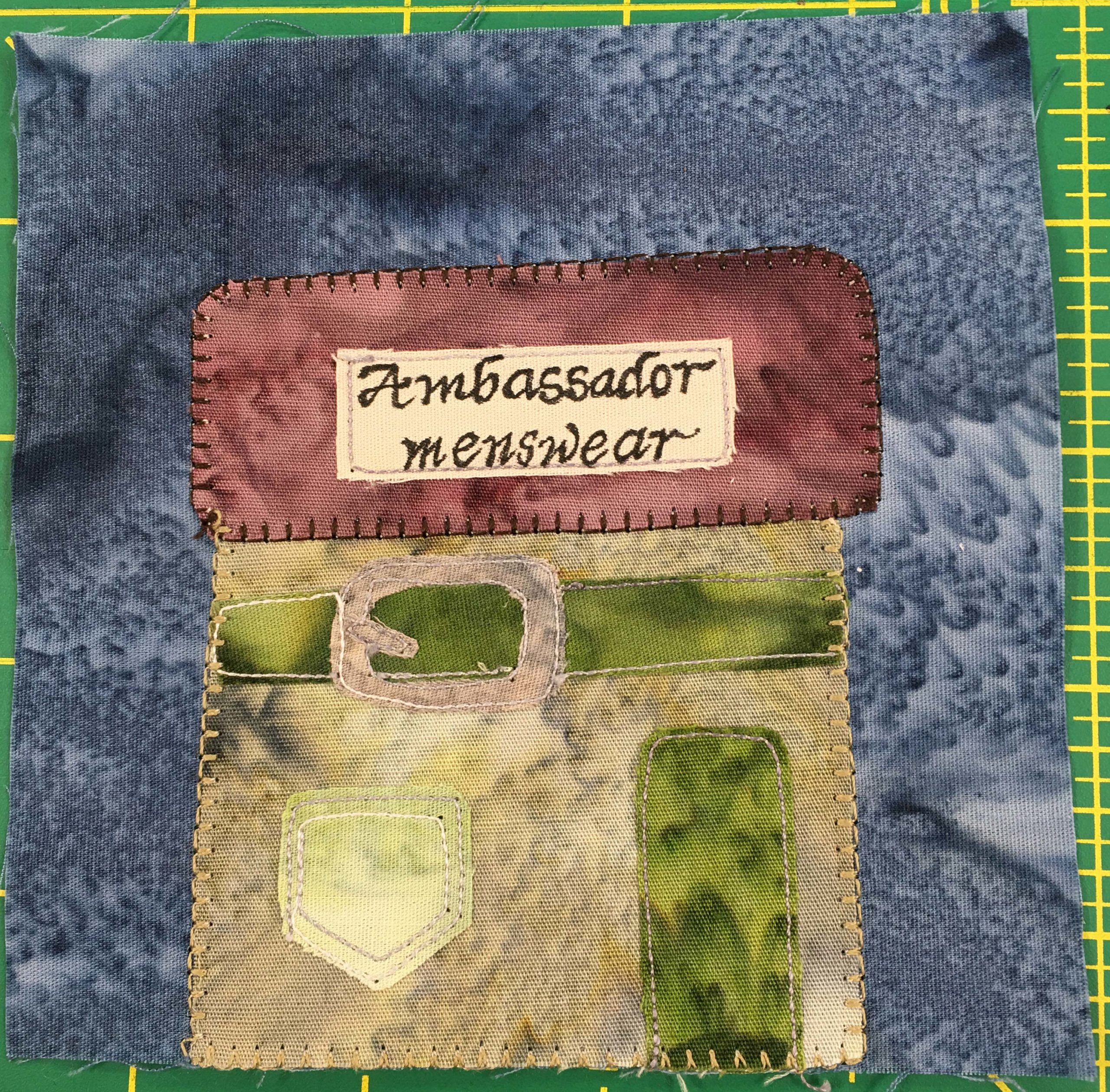 This quilt block shows a square building designed to look like clothing. It has a large green belt and buckle under the roof, and a window shaped like a pocket you might find on pants or kilt. There is a green door to the right. The purple square roof has a sign that reads, 