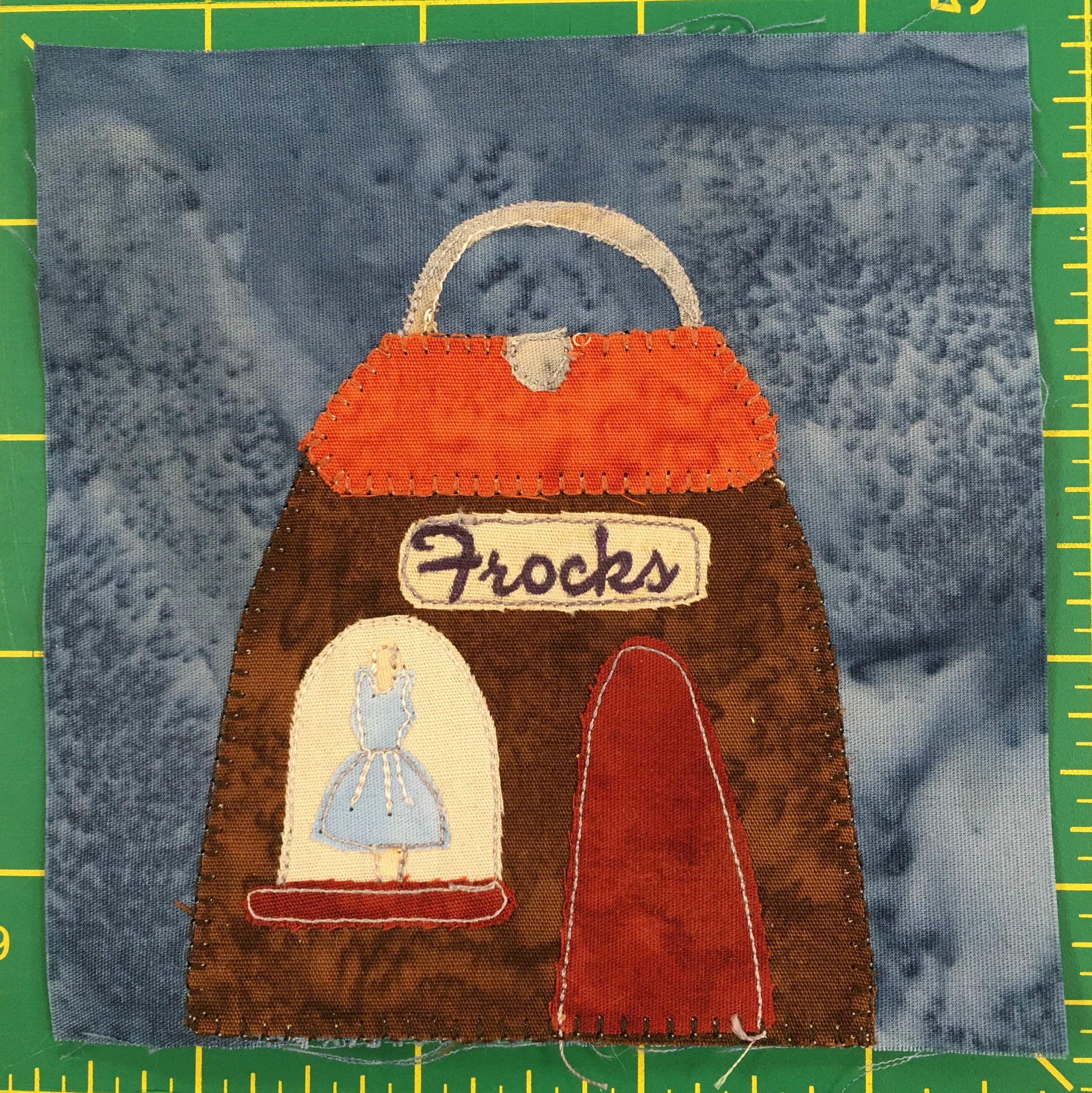 This quilt block shows a building shaped like a handbag. It is brown with an orange flap roof, a gray clasp and a gray handle. In the display window on the left of the building is a blue dres, and to the right of that a red door. There is a sign on the front that reads,