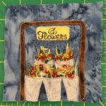 This quilt block shows a flower stand: lots of baskets of flowers encased in a brown square bracket with a sign on top that reads,