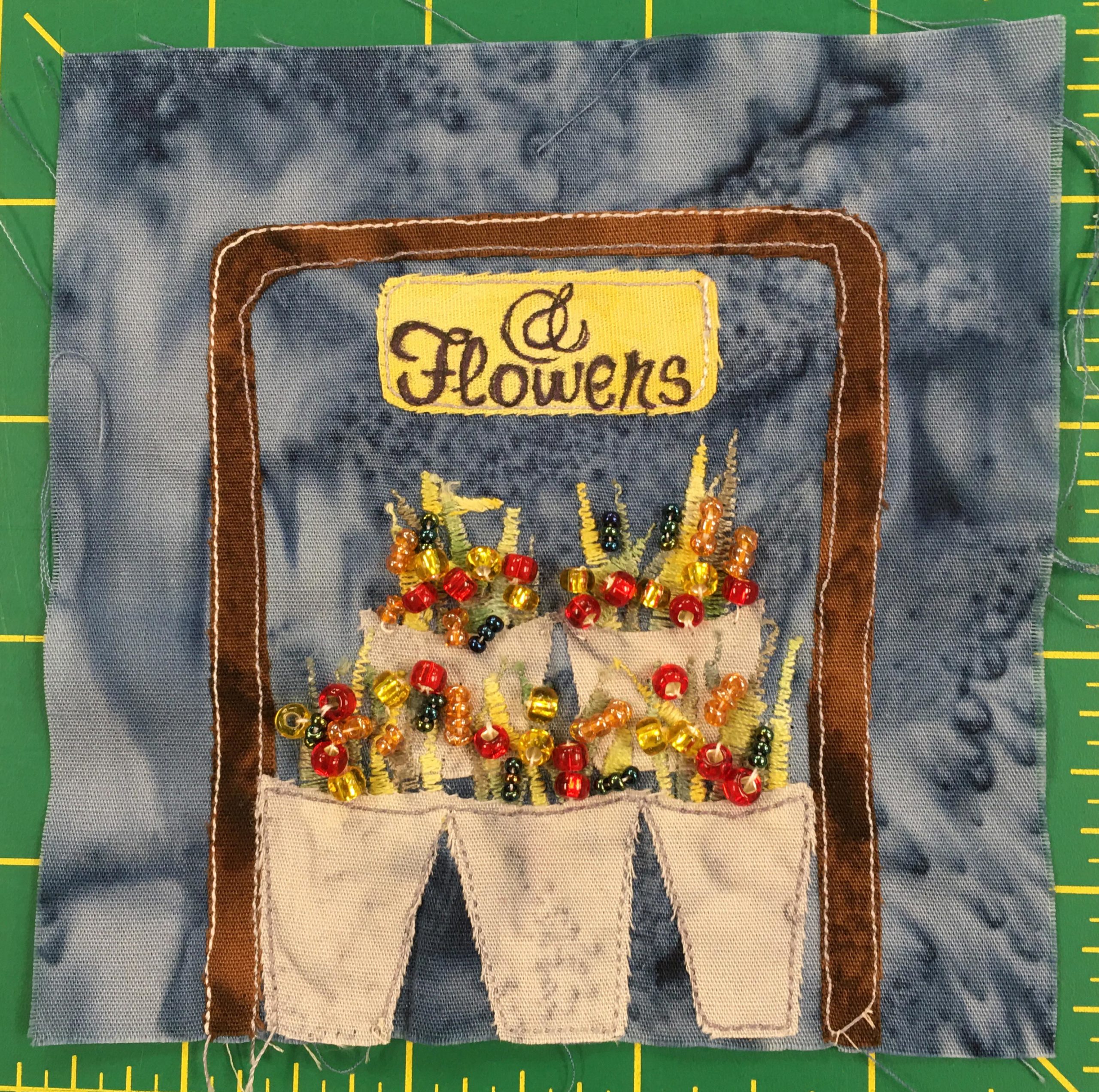 This quilt block shows a flower stand: lots of baskets of flowers encased in a brown square bracket with a sign on top that reads,