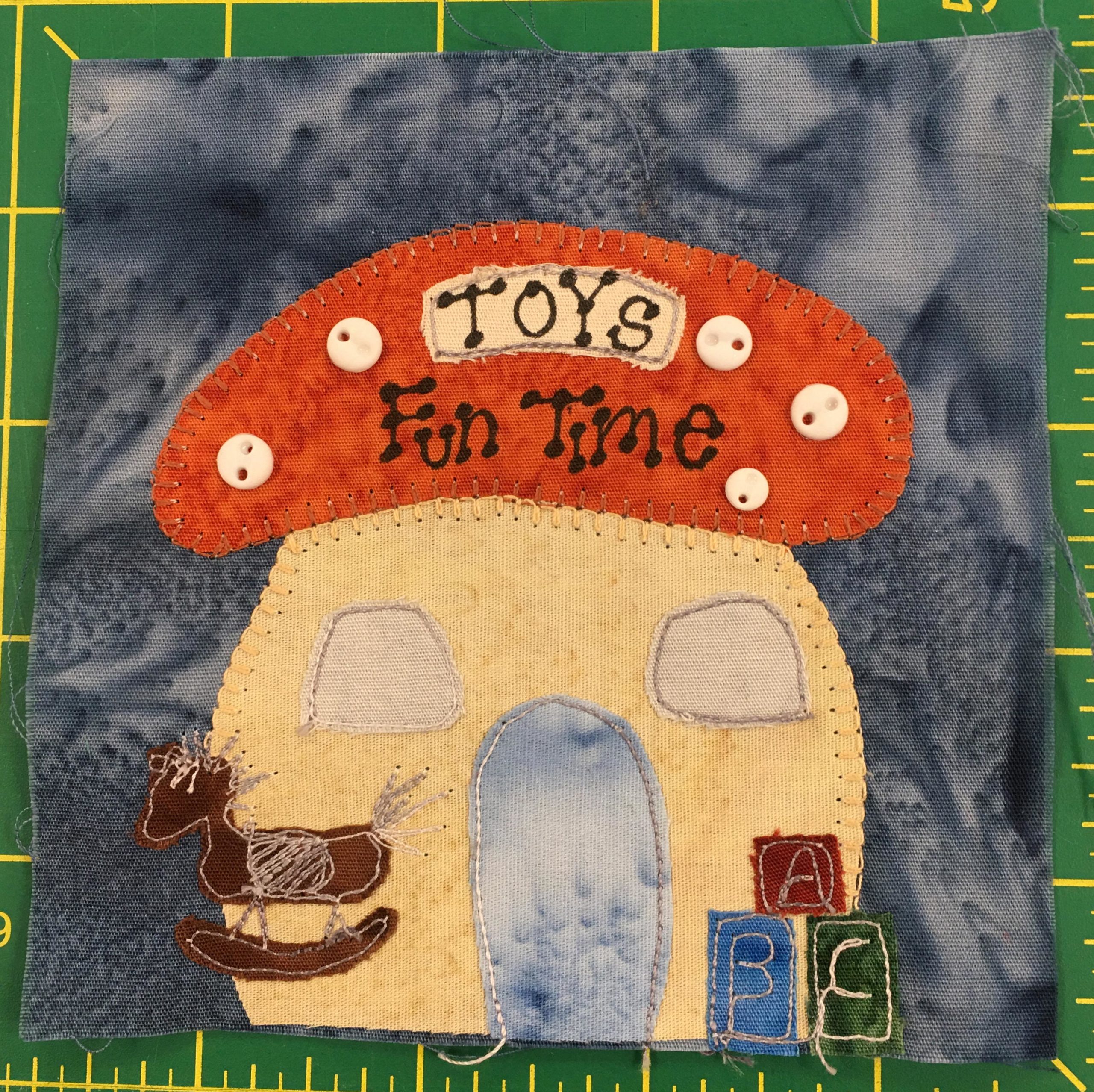 This quilt block shows a mushroom shaped building with an orange polka-dotted top. The polka-dots are made with little light two-holed buttons. On the cap roof it reads, 