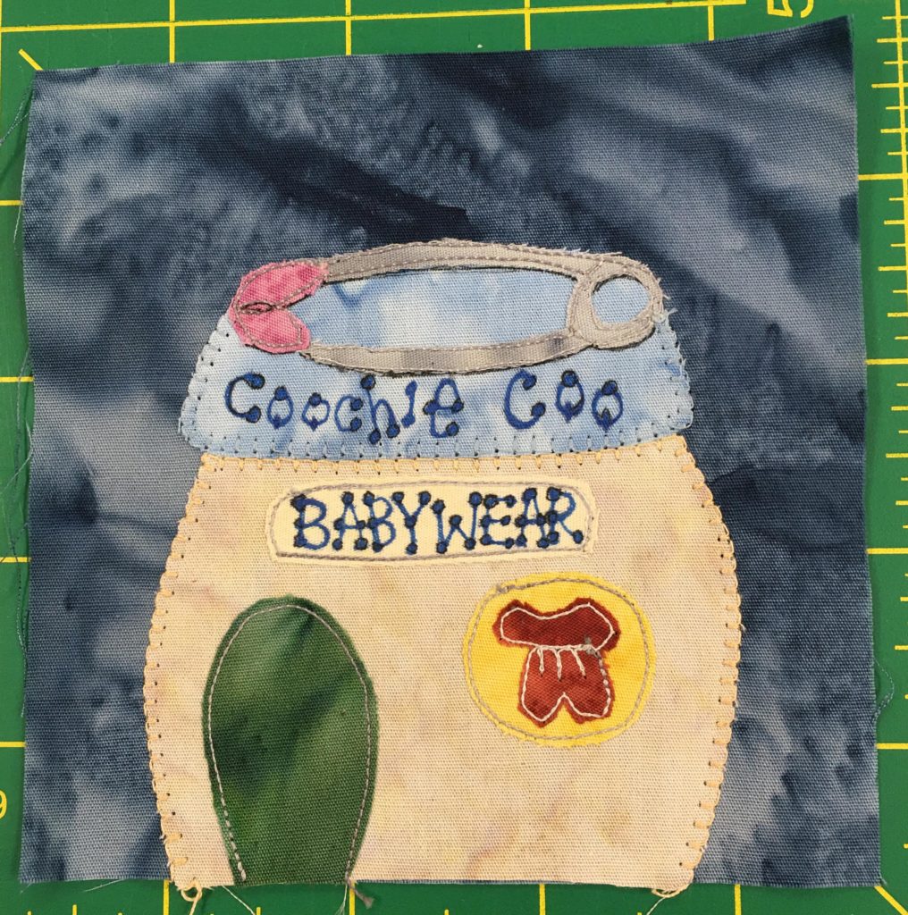 This quilt block shows a building shaped a little bit like the lower part of a baby bottle. It is plump and wide with a cap-like baby-blue roof. On the roof is a giant diaper safety-pin that is pink on the left end. Letters on the building read,
