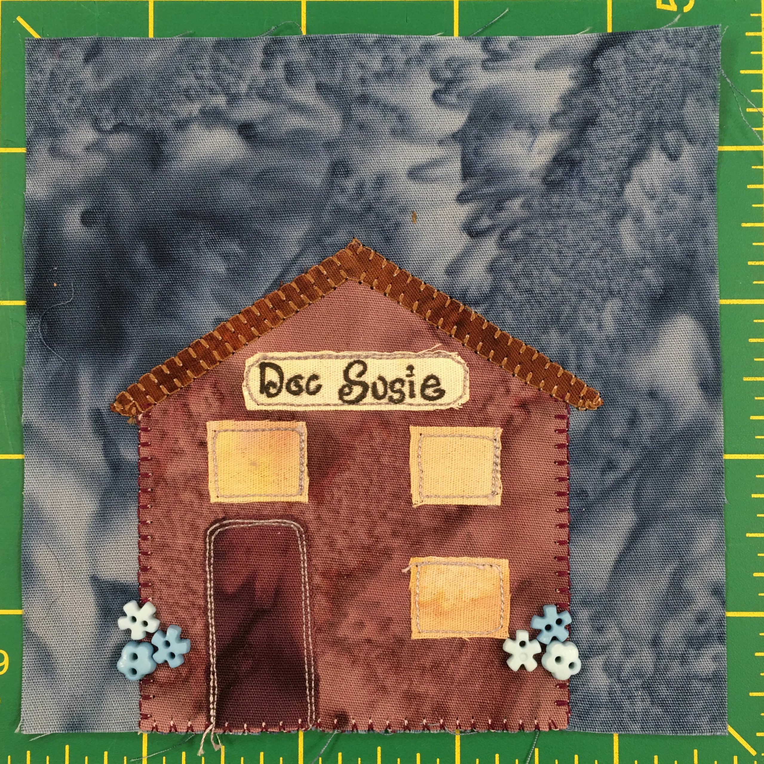 This quilt block shows a brown building with three golden windows. Two vertically on the right side, and one above the darker brown door. The roof is a simple angled roof. Under the rood if a sign in cursive that says,