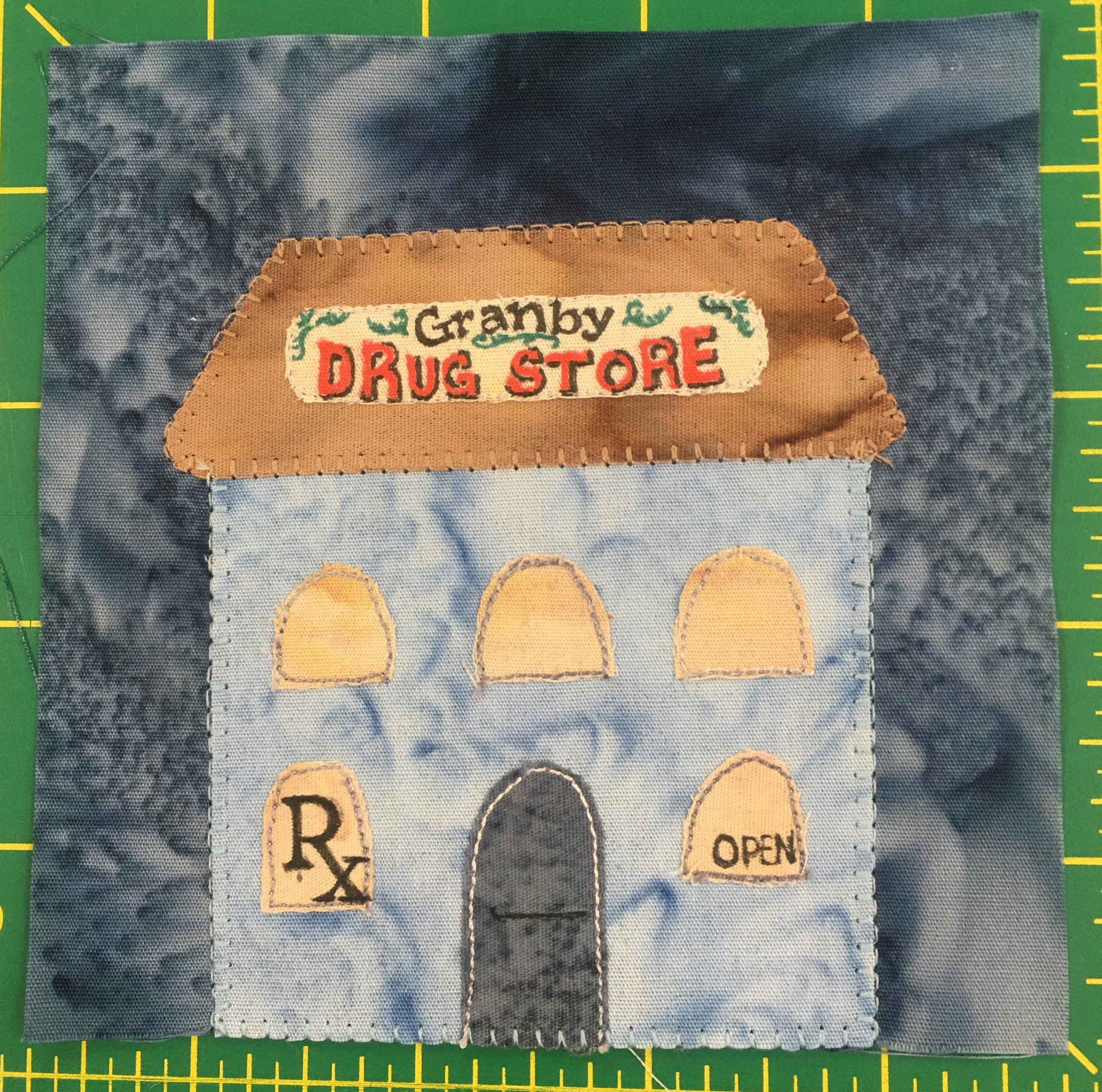 This quilt block shows a tall building with a trapezoid shaped orange roof that has a sign on the front reading,