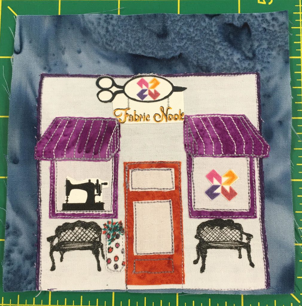 This quilt block shows the front of the Fabric Nook store, complete with the logo at the top of the building. It is a square all white building with no separate roof. There are two windows on either side of the center door with purple canopies. On the left is a silhouette of an old school sewing machine, and on the right is the Fabric Nook quilt block logo. There are two ornate benches in front of the store, as well as a planter that is decorated with polka dots. The door is orange with two sections for glass.