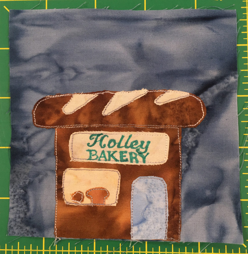 This quilt block shows a brown building with a roof in the shape of a bread loaf. The sign on the front reads,