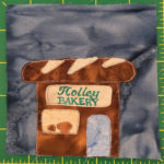 This quilt block shows a brown building with a roof in the shape of a bread loaf. The sign on the front reads, 