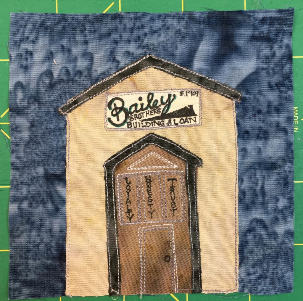This quilt block shows a tall intimidating building with a large section in the front that looks like a door. On the faux brown door is some trim and in the trim are the words,