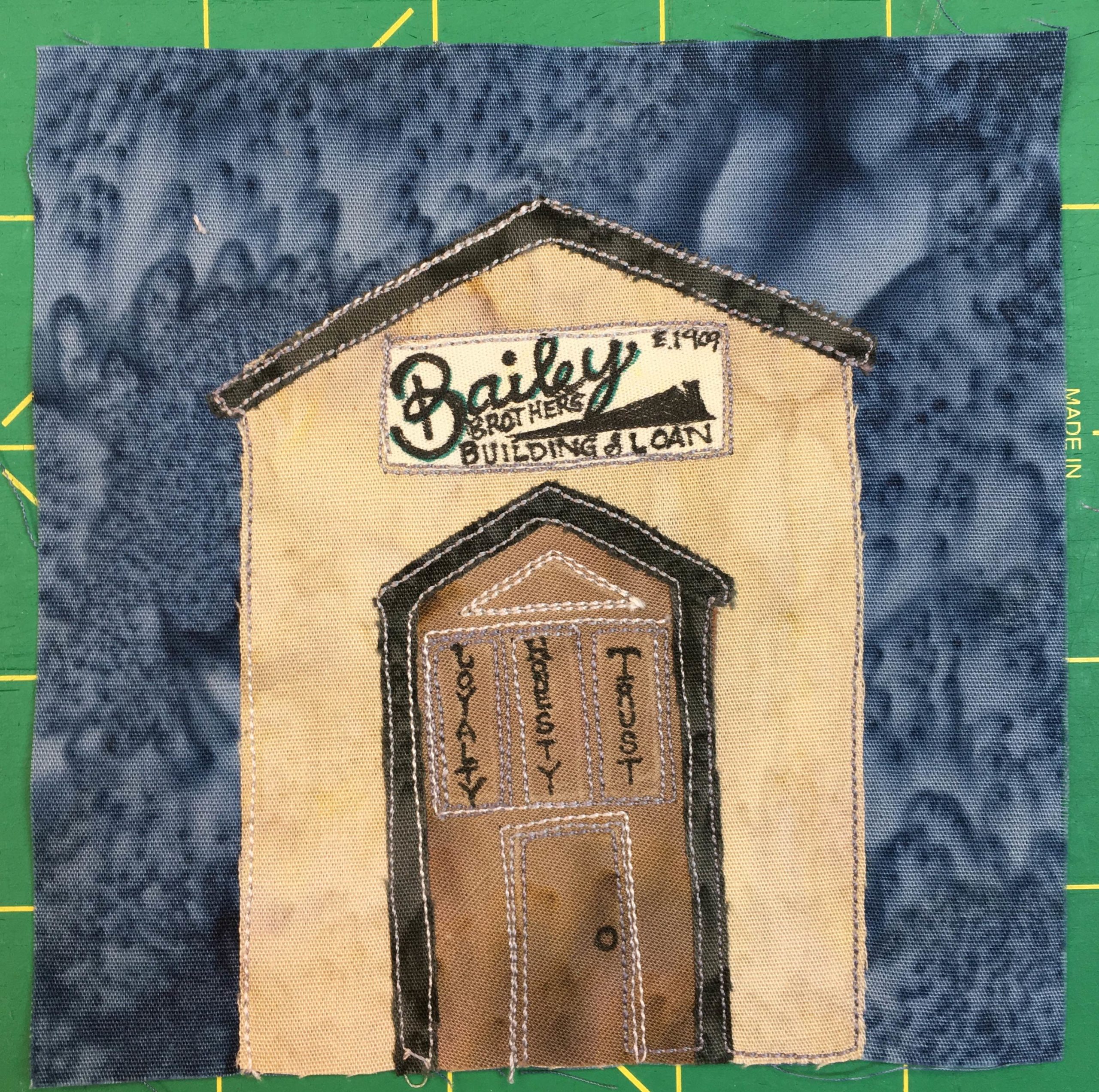 This quilt block shows a tall intimidating building with a large section in the front that looks like a door. On the faux brown door is some trim and in the trim are the words, 