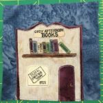 This quilt block shows a store front of a bookstore. The building's decorative roof is shaped in such a way that it makes the building look like an open book. Under the roof lining is a sign that says,