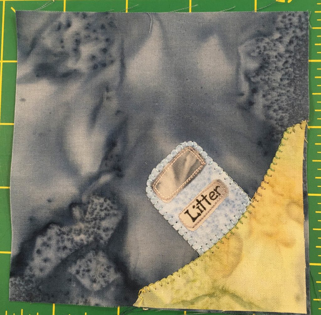 This quilt block shows a corner with a yellow-green patch of ground in the bottom right. On that patch of ground is a blueish-gray public box with a flap on it and the word