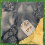 This quilt block shows a corner with a yellow-green patch of ground in the bottom right. On that patch of ground is a blueish-gray public box with a flap on it and the word