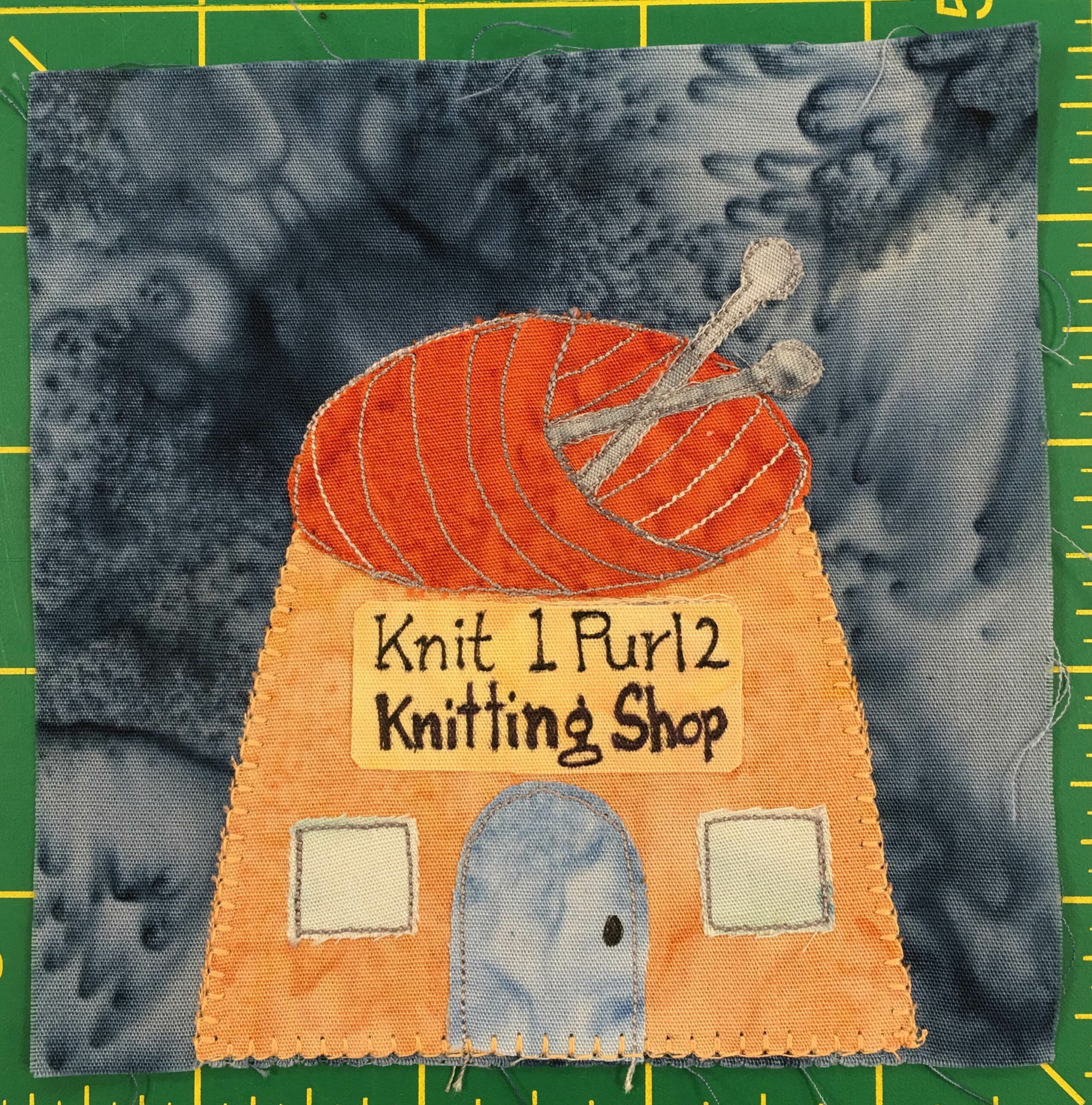 This quilt block shows a somewhat squat building whose roof looks like a ball of orange yarn with knitting needles poked into it. Below the orange yarn is a light orange exterior with a big yellow sign on the front hat says, 