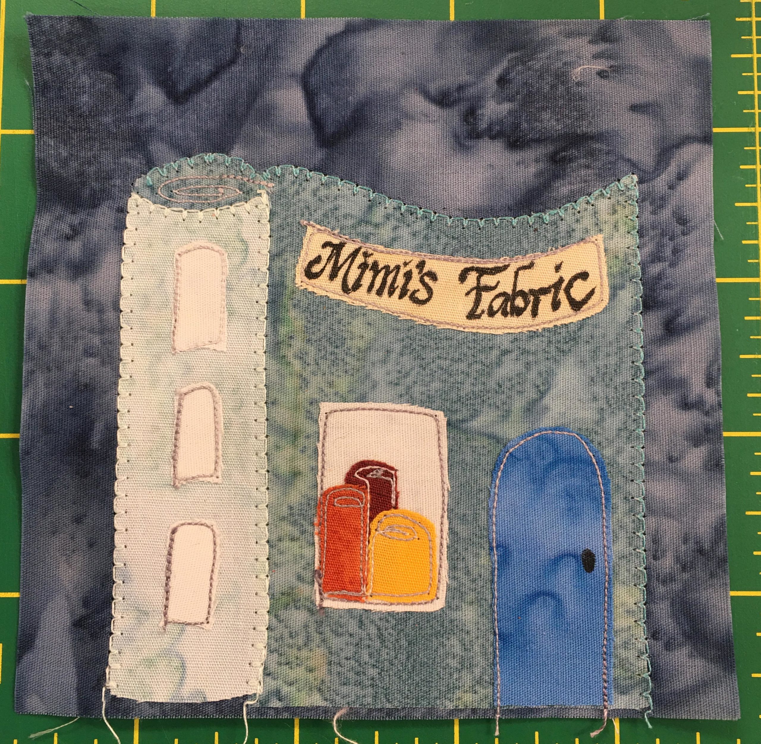 This quilt block shows a very interesting building that's in the shape of a roll of fabric! It's unrolls a bit giving the building a front. There's a blue door on the right, and a window to the left of it with trio of colored fabric rolls inside. There are three windows going up the roll part of the building. Across the top of the 