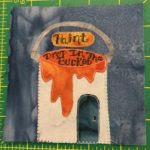 This quilt block shows a building shaped like a paint bucket. The paint that is glazing over the side of the can is orange. The building or can is white, and there's a semi-circle handle over the roof. The sign above the open hole in the bucket reads,