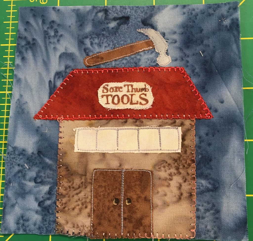This quilt block shows a square brown building with a rhombus shaped rust colored roof. On top of the roof is a giant sign in the shape of a hammer, while on the roof itself is a sign that reads,