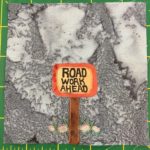 This quilt block shows a lighter background color than previous blocks. A single sign with some rocks behind it stands at the top of a wooden post in this block. An orange border is around a yellow square with the words inside saying,