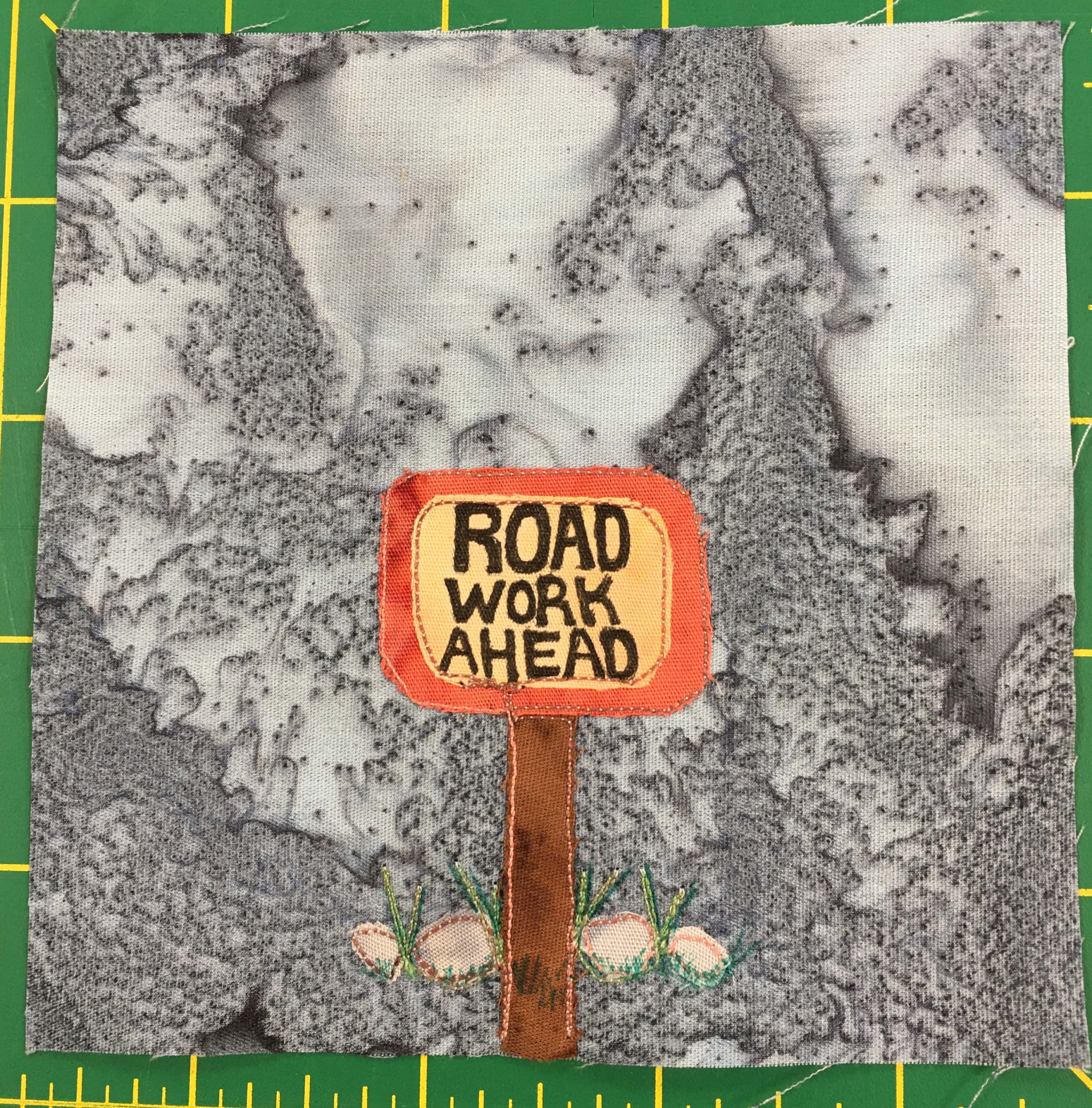 This quilt block shows a lighter background color than previous blocks. A single sign with some rocks behind it stands at the top of a wooden post in this block. An orange border is around a yellow square with the words inside saying, 